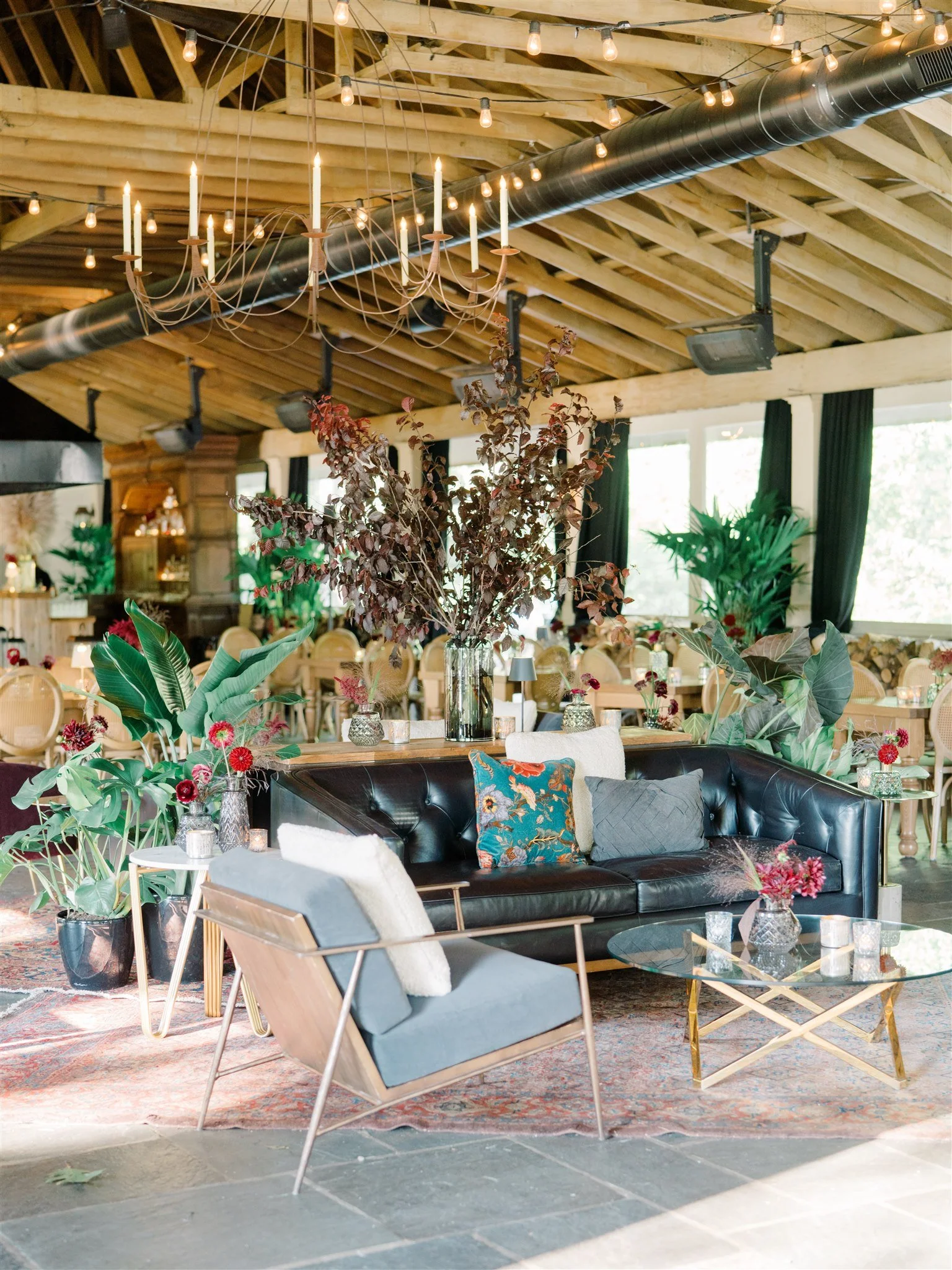 A stylish lounge area with a black leather couch, colorful pillows, modern chair, glass coffee table, large glass vase with dark red and purple branches, lush green plants, candle centerpieces, and rustic wooden ceiling with string lights and a chand