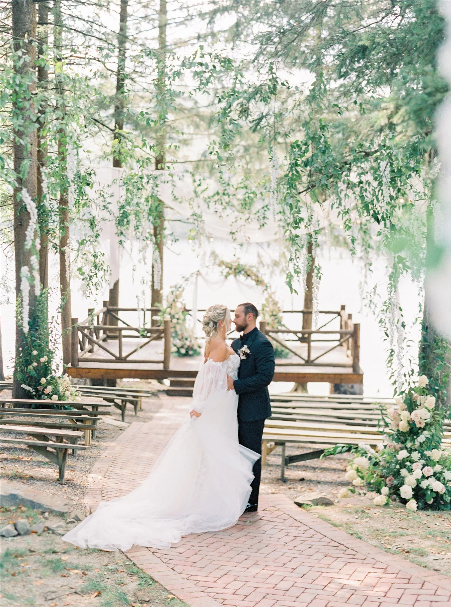 A bride and groom are standing close together in a wooded outdoor wedding setting, about to kiss. The bride is in a white gown with off-the-shoulder sleeves, and the groom is in a dark suit. They are surrounded by trees, floral arrangements, and wood