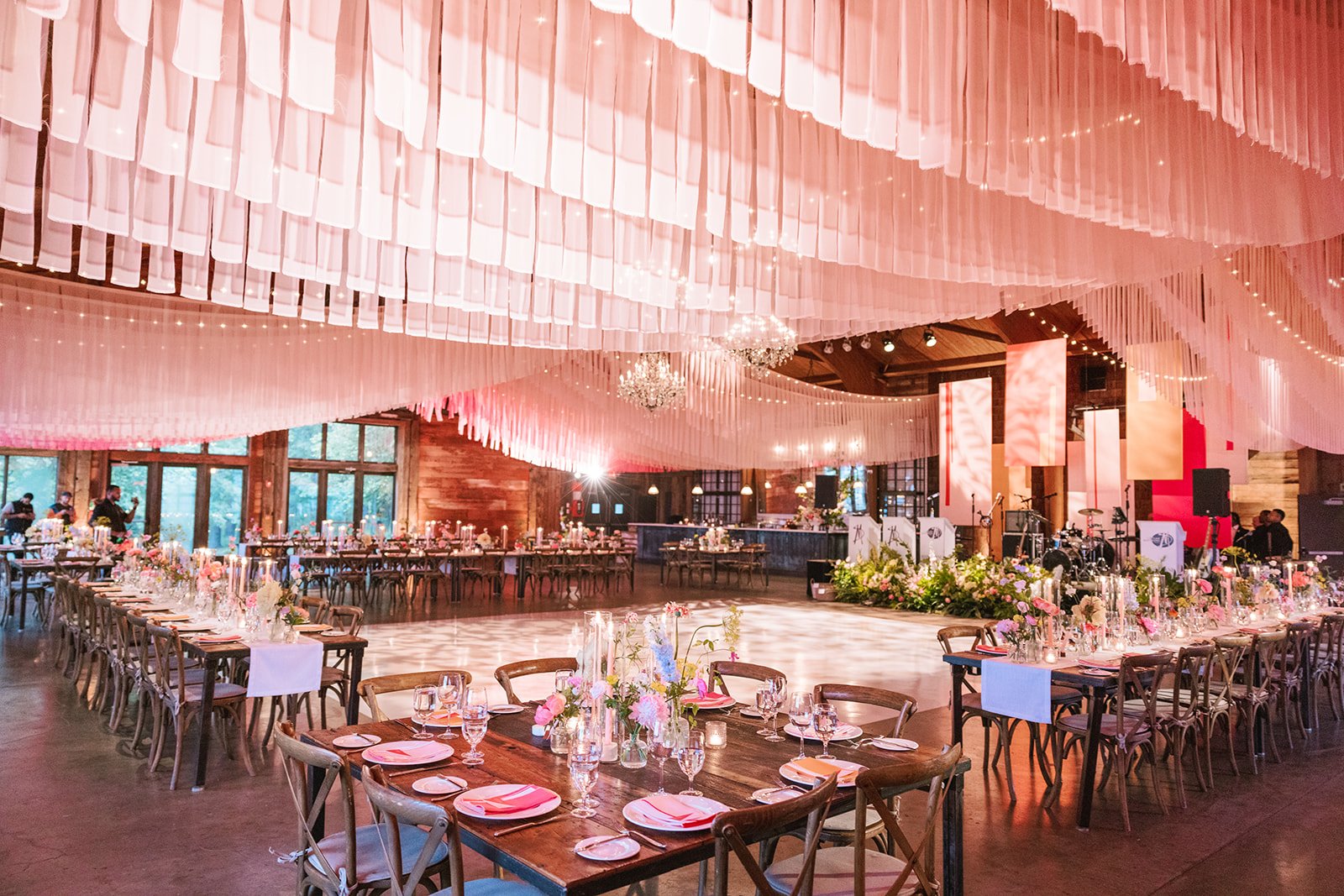 Elegant event hall decorated with pink drapery and string lights, featuring arranged tables with floral centerpieces and place settings, a dance floor in the center, and a stage with musical equipment in the background.
