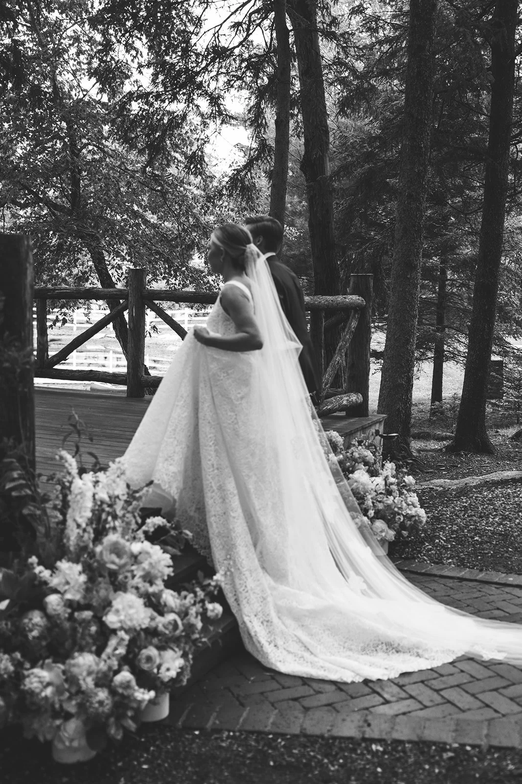 A bride and groom standing together outdoors on a wedding day, with the bride in a white wedding dress and veil, and the groom in a dark suit. They are near a wooden fence and surrounded by trees and floral arrangements.