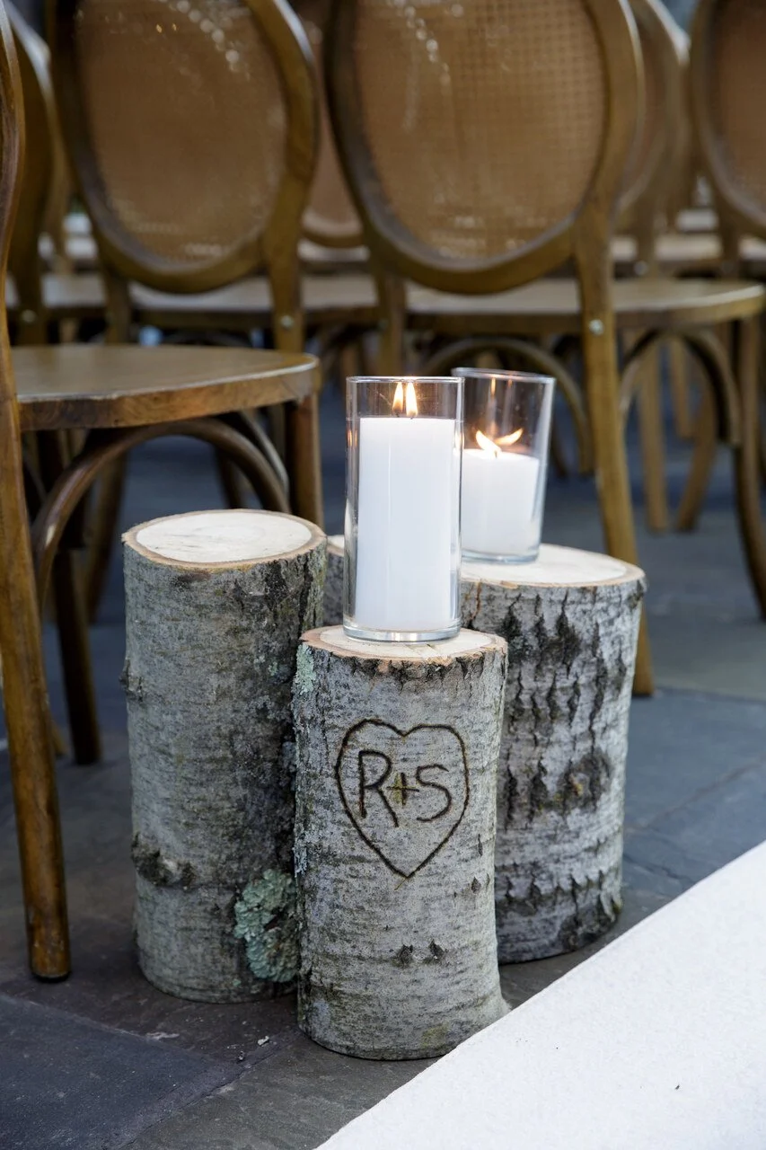 Three birch logs with candles and a heart carving engraved with 'R + S', surrounded by wooden chairs.