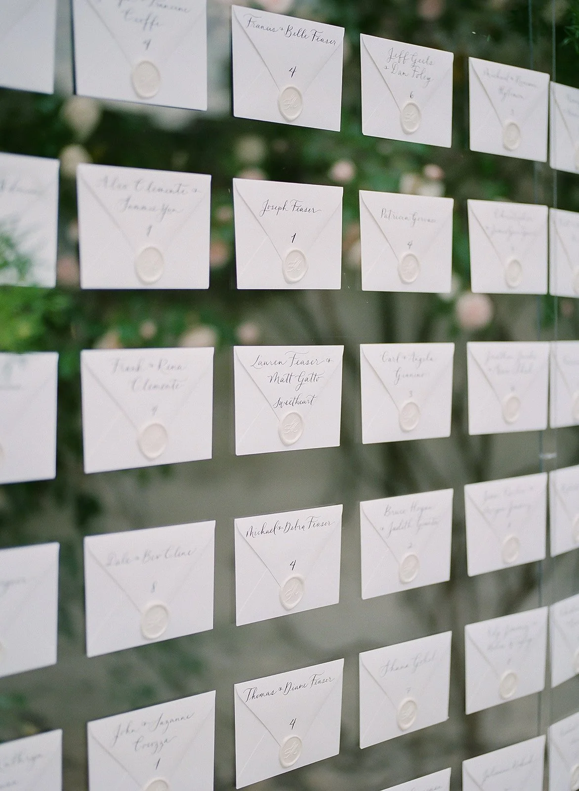 A display of white wedding place cards arranged on a glass or acrylic board, each sealed with a wax emblem, with handwritten names and table numbers visible, set against a blurred outdoor background.