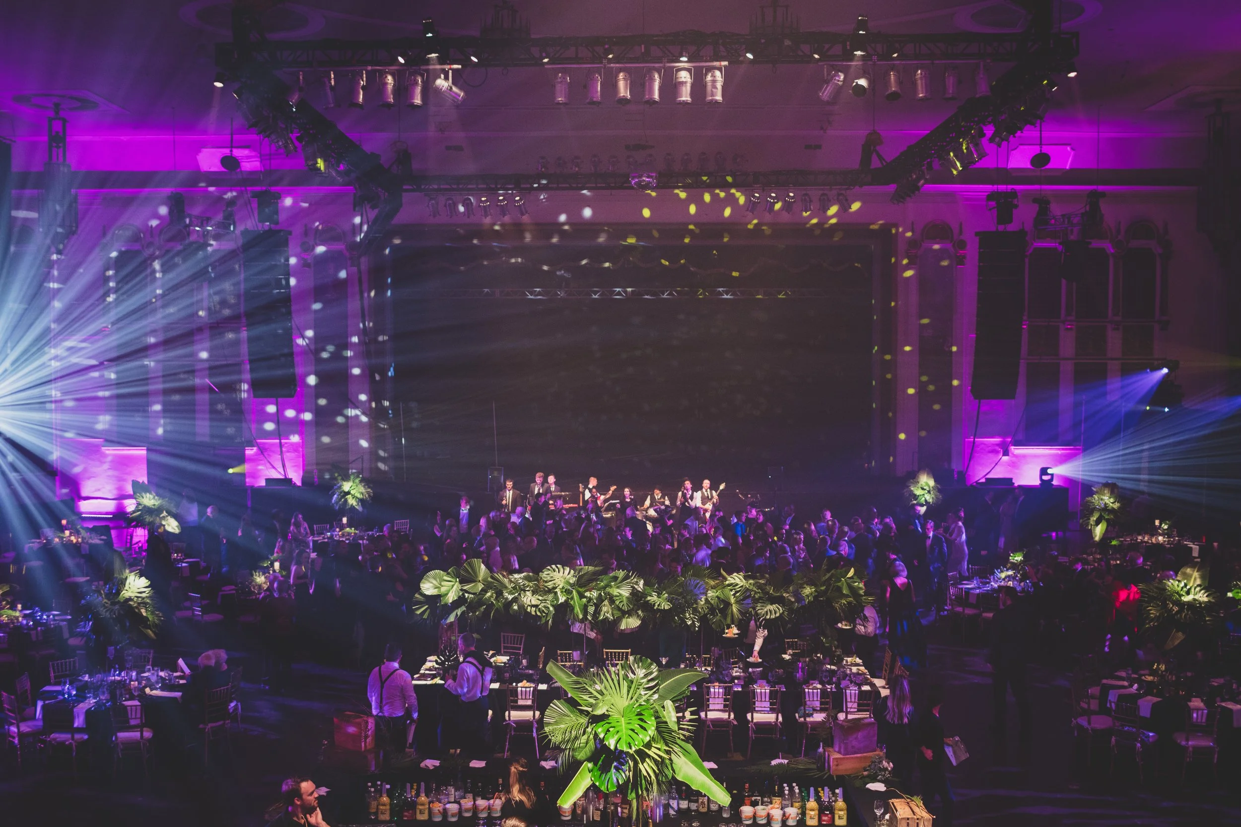 An indoor event with purple and yellow stage lighting, a live band performing on stage, and a large audience. The venue is decorated with green tropical plants and has tables set up for dining.