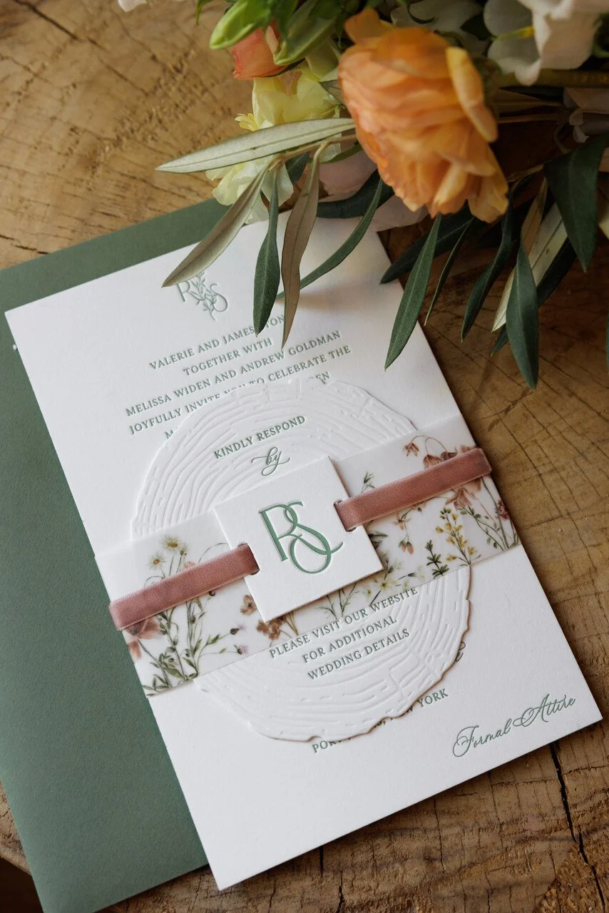 Invitation card with floral design and embossed details, ribbon, and a monogram with the initials R and S, placed on a wooden surface along with a bouquet of flowers.