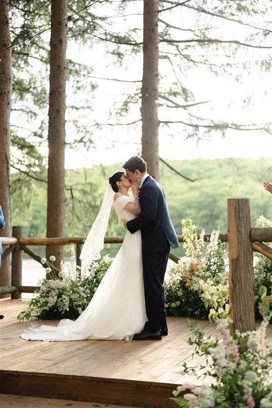 A bride and groom share a kiss during their outdoor wedding ceremony, surrounded by trees and floral decorations.