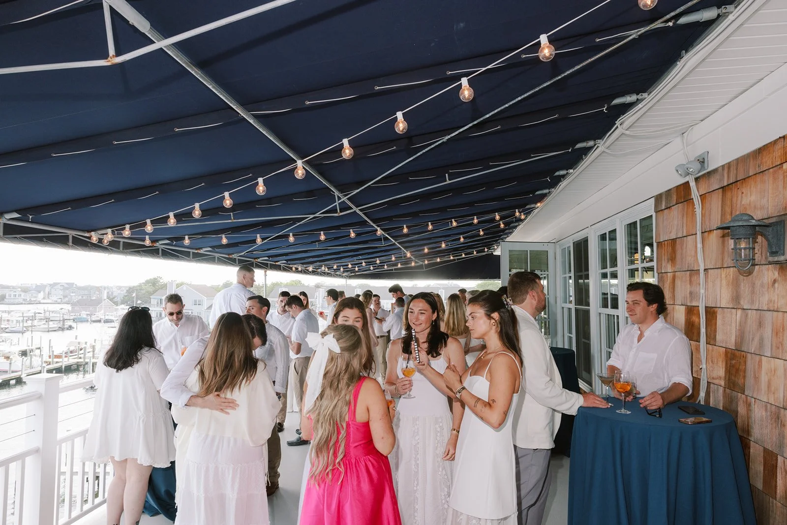 People at a social gathering on a decorated outdoor patio with hanging string lights, overlooking a marina.