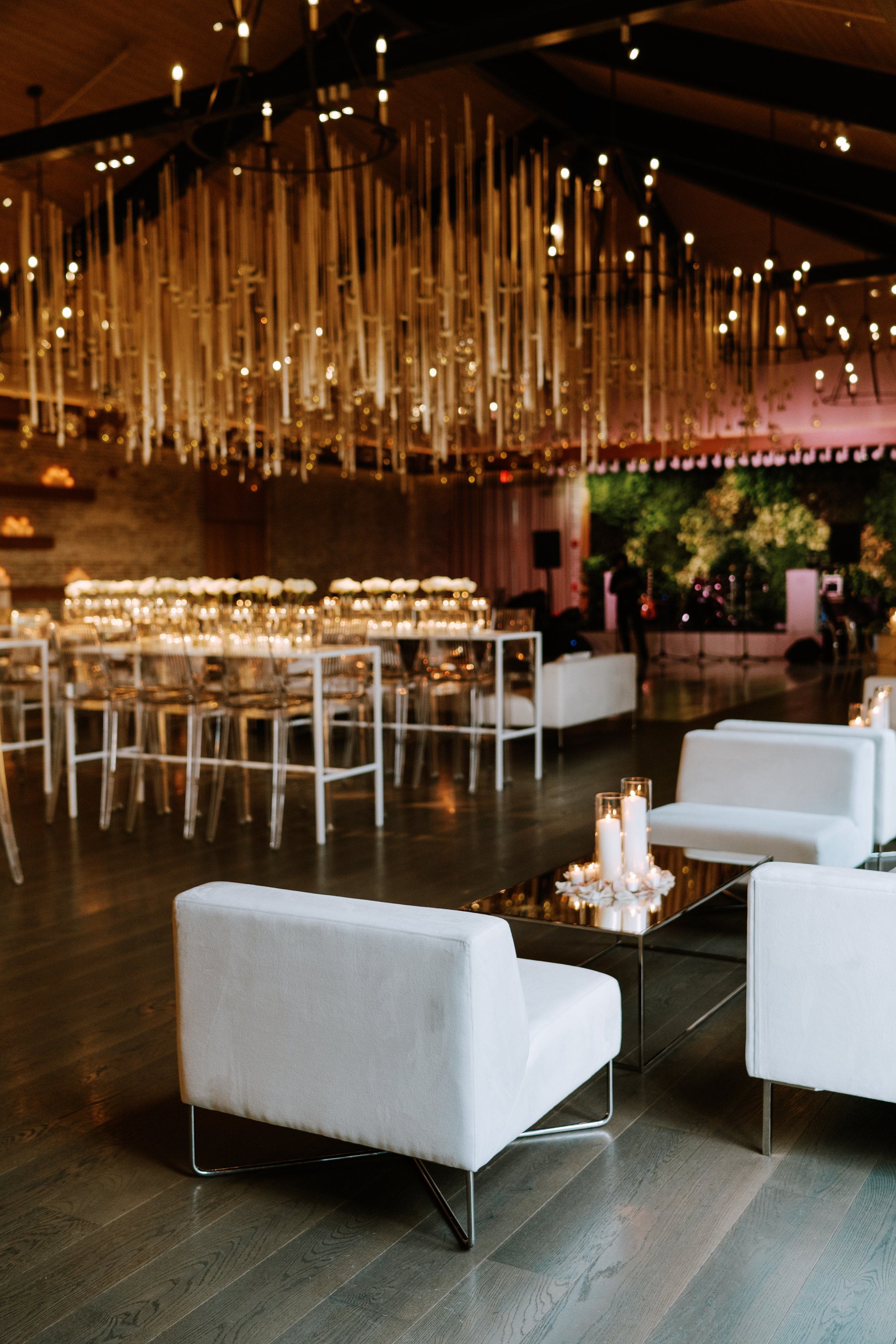 Elegant event space with white armchairs, candles, and modern decor, featuring hanging lights and a long dining table set for a formal occasion.