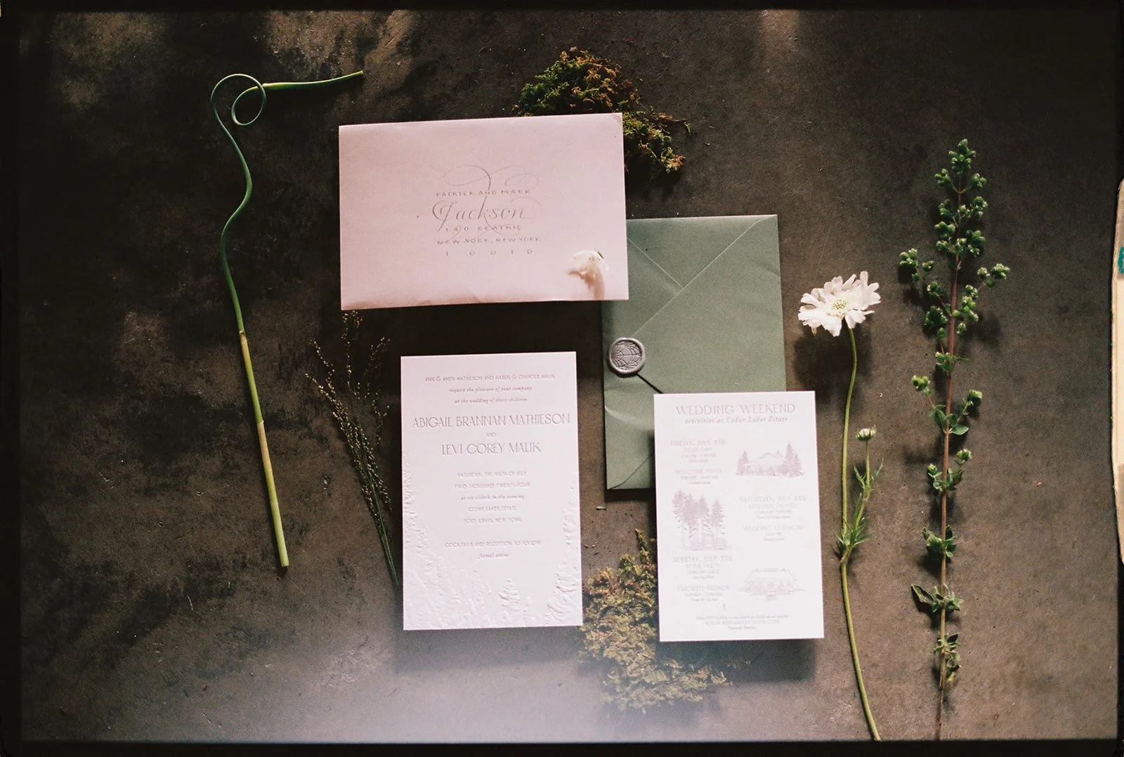 Wedding invitation set with green envelope, printed cards, and decorative flowers on a dark surface.
