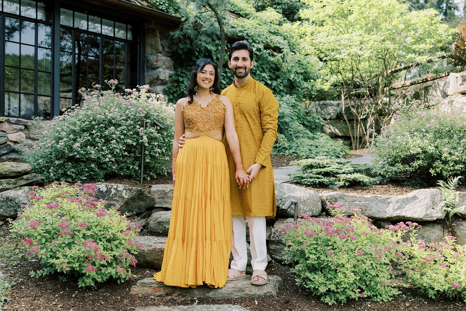 A man and a woman stand together outdoors in a garden, holding hands and smiling. The woman is wearing a yellow dress with intricate gold embroidery on the bodice, and the man is wearing a traditional mustard yellow kurta with white pants. The backgr