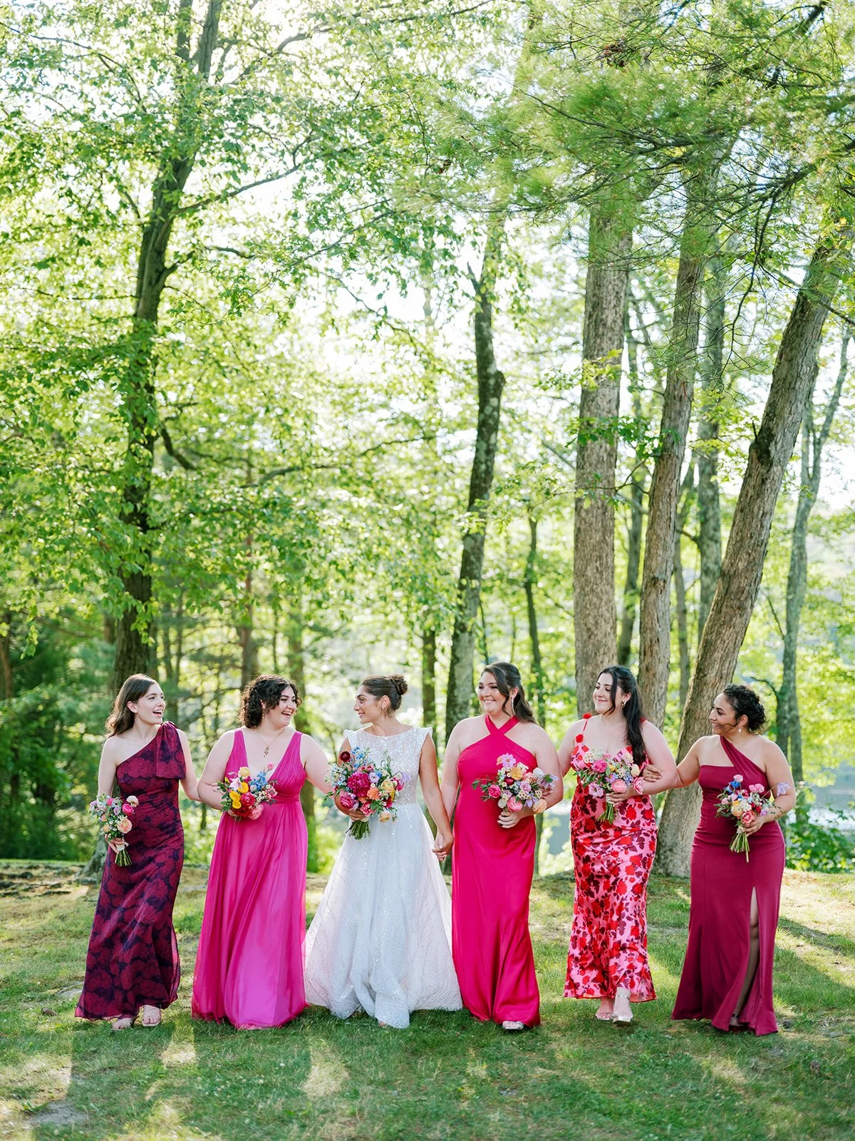 A bride and five bridesmaids walking and holding bouquets in a wooded outdoor setting with green trees and sunlight.