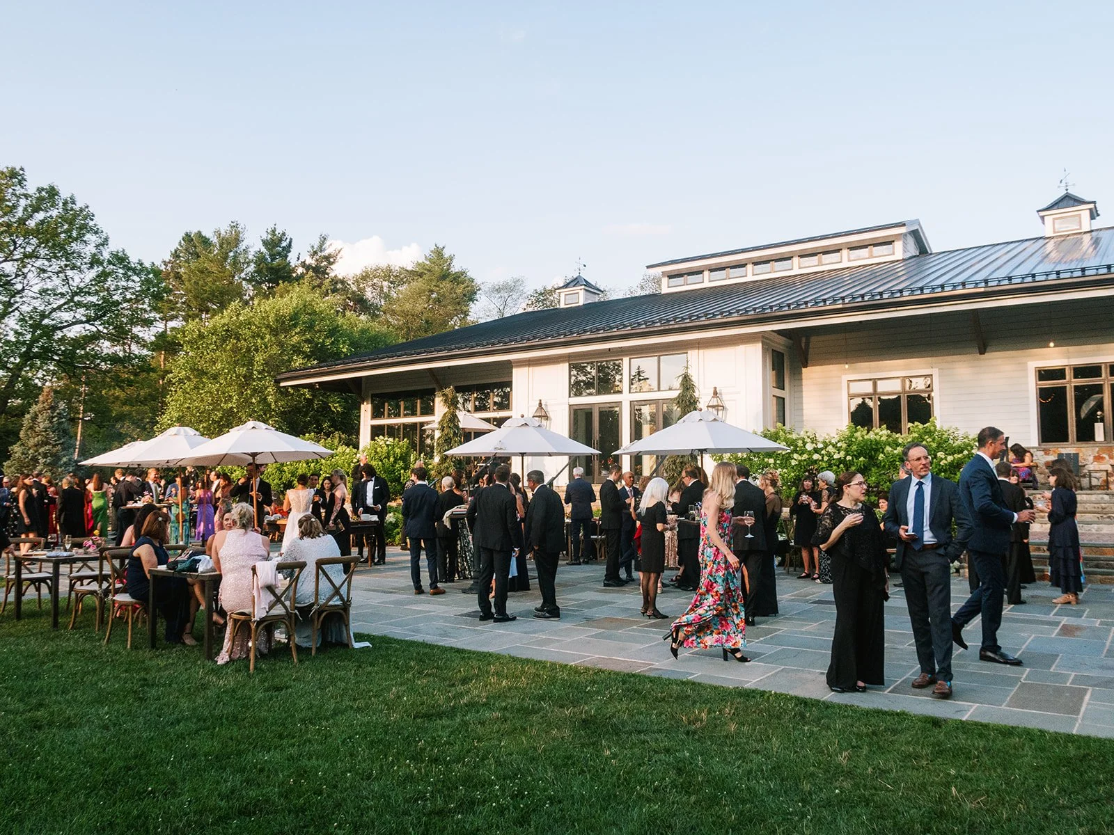 Guests at an outdoor wedding reception gather on a patio with white umbrellas, dressed in formal attire, with a large house or event venue in the background surrounded by greenery and trees.