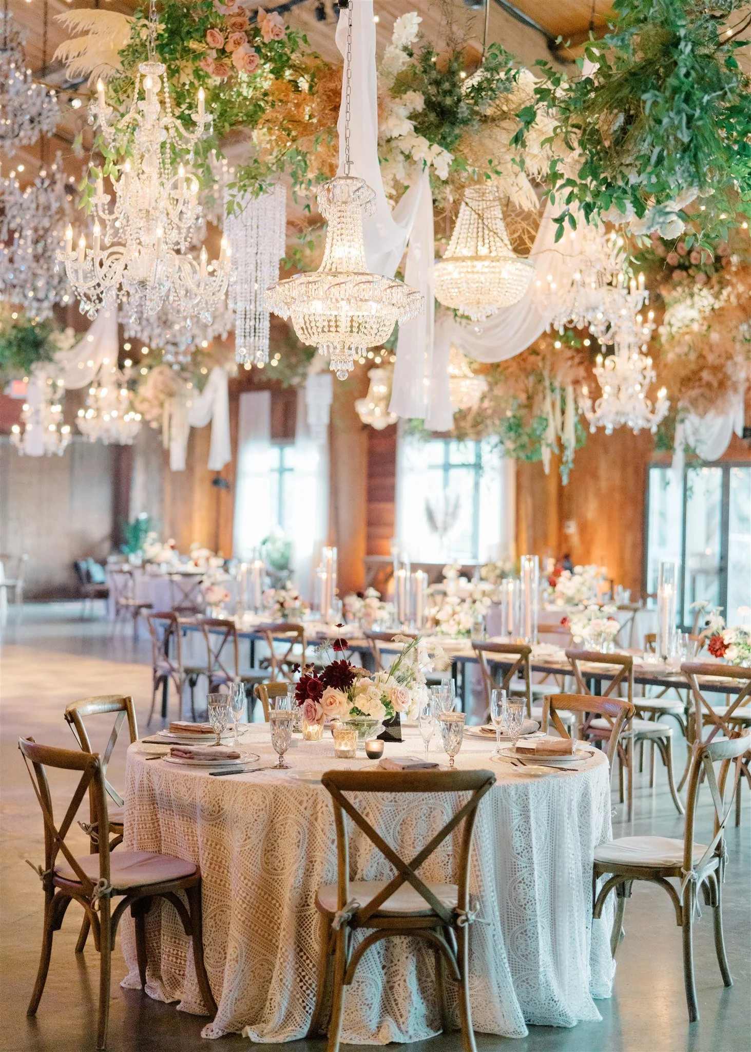 An elegant event space decorated with chandeliers, floral arrangements, and draped fabric. Round tables with lace tablecloths are set with glassware, plates, and floral centerpieces. The room has large windows and wooden walls, creating a warm, invit