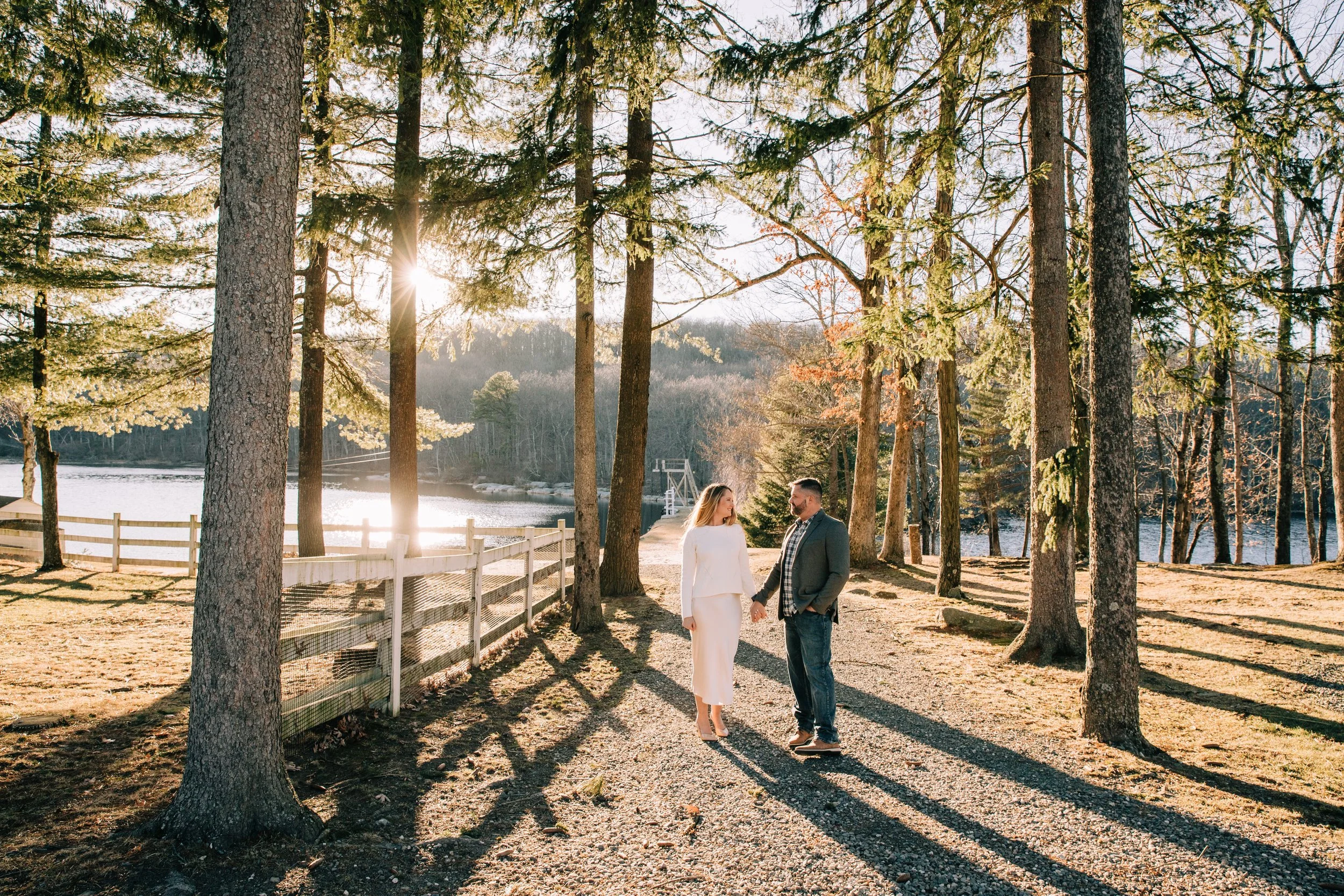 A couple holding hands and smiling at each other outdoors in a wooded area by a lake, with sunlight shining through the trees, during what appears to be late afternoon or early evening.