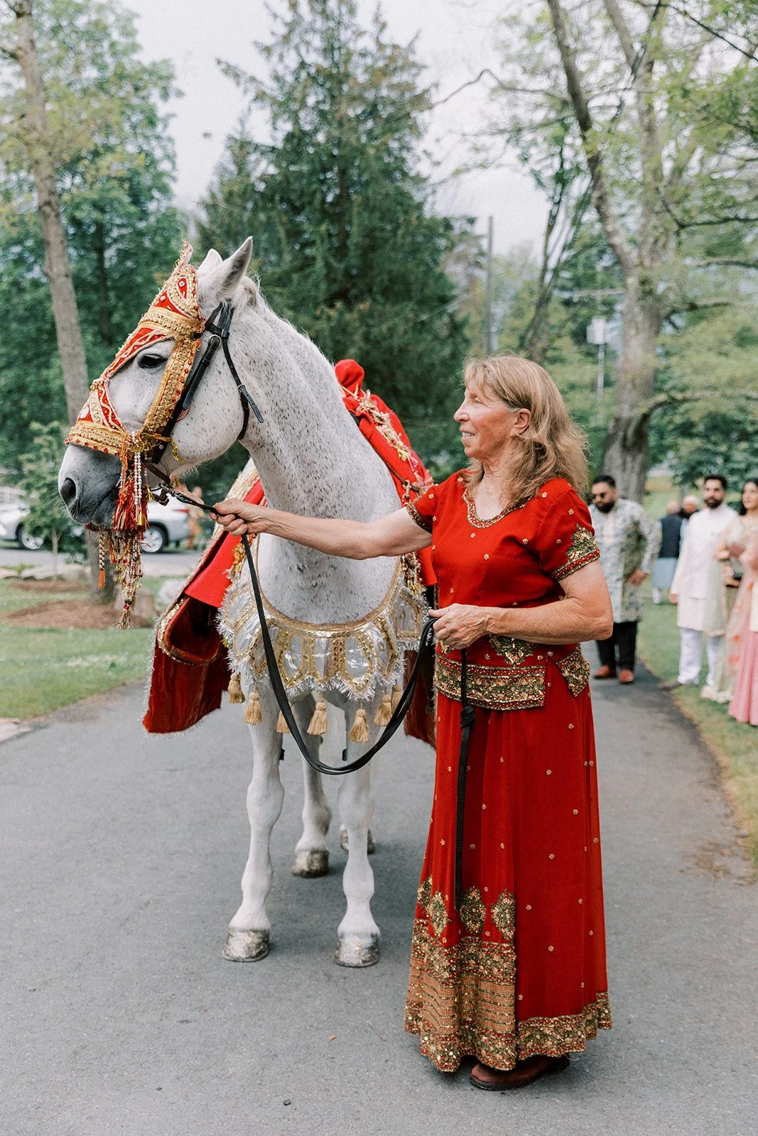 A woman in a red traditional dress standing next to a decorated white horse during a festive outdoor event with people in the background.