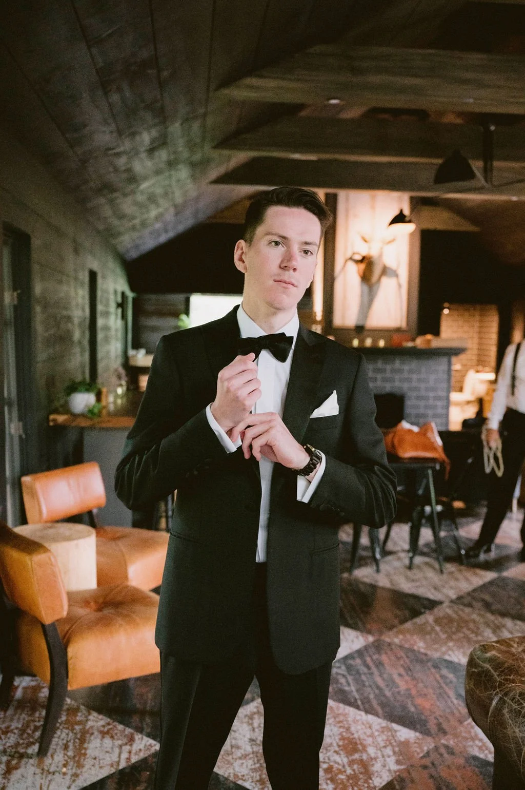 Young man in a black tuxedo with a bow tie, adjusting his bow tie, standing in a warmly lit indoor space with rustic decor.