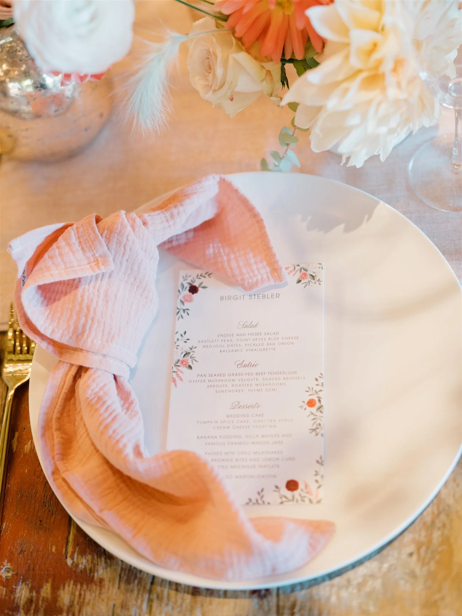 A floral centerpiece with white and peach-colored flowers and some greenery on a wooden table. A white plate holding a pink napkin and a printed menu with floral borders is also on the table.