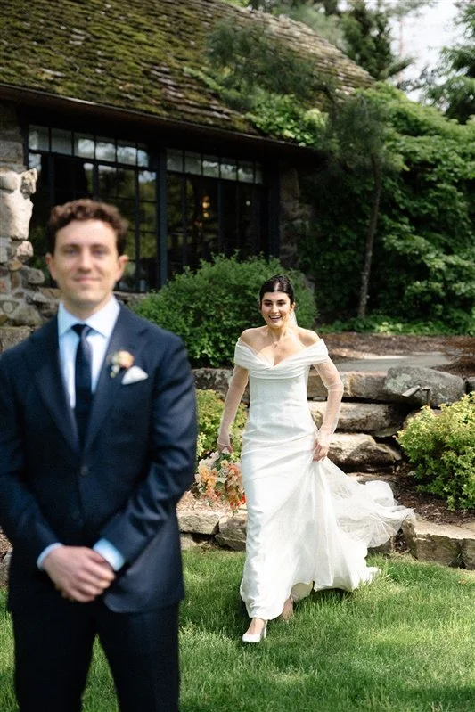 A bride in a white wedding dress holding a bouquet and a groom in a suit, with a garden and stone building in the background.