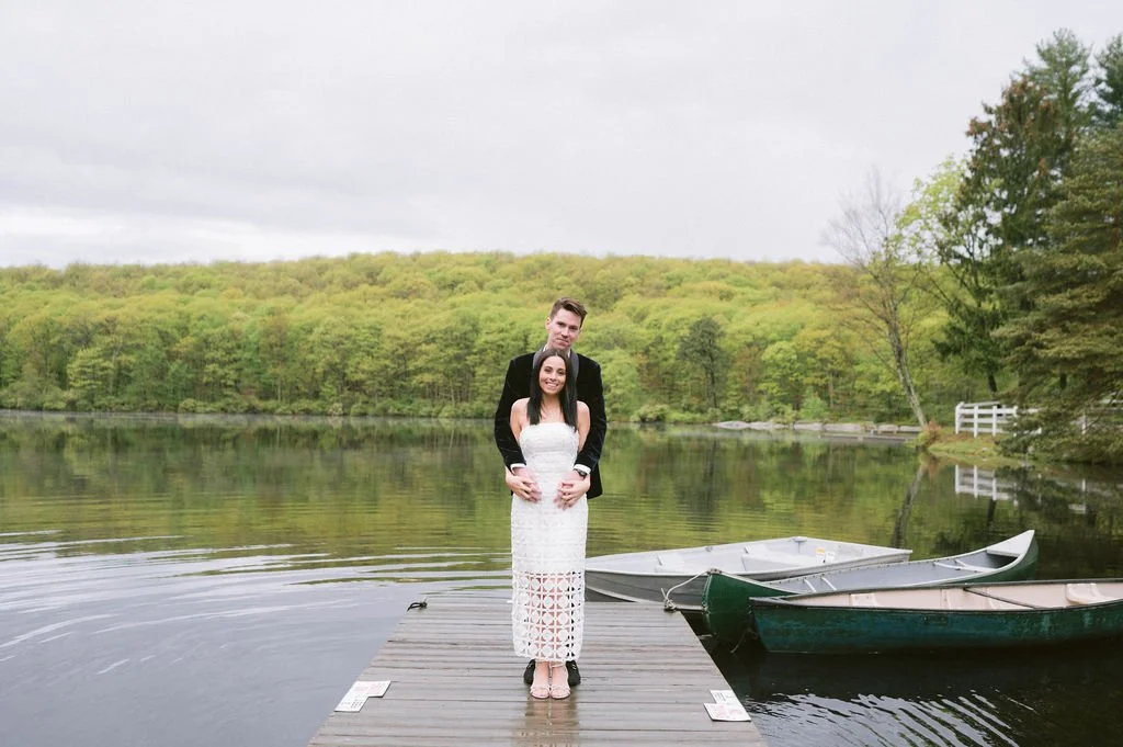 A couple standing on a dock by a lake, with lush green trees in the background, and two rowboats moored nearby.