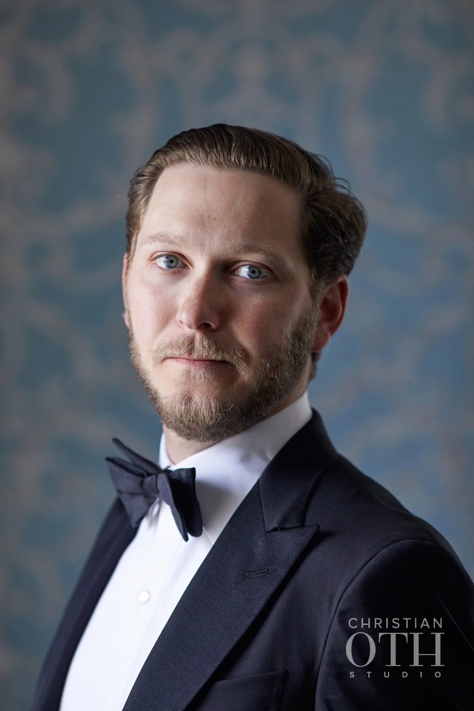 A man wearing a tuxedo with a bowtie posing for a portrait in front of a textured blue background.