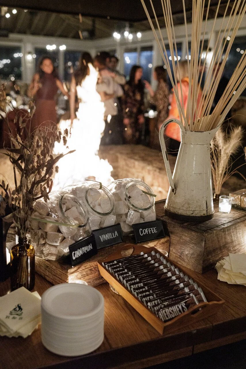 Table with marshmallows labeled 'cinnamon,' 'vanilla,' and 'coffee,' a pitcher with skewers, a tray of Hershey's chocolates, a stack of plates, and a background with blurred people at a social gathering.