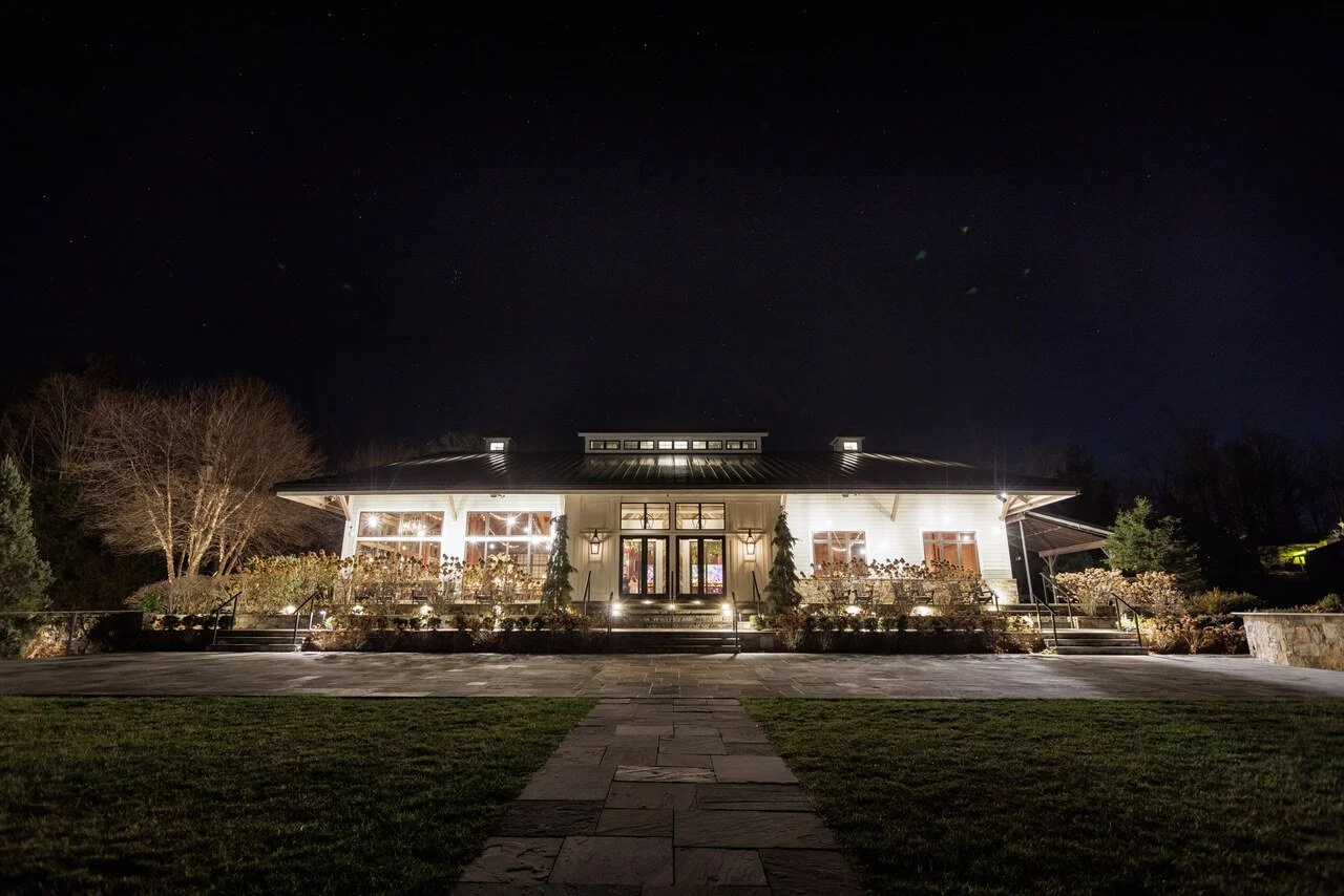 Night view of a well-lit house with a metal roof and large front porch, surrounded by landscaping, under a starry sky.