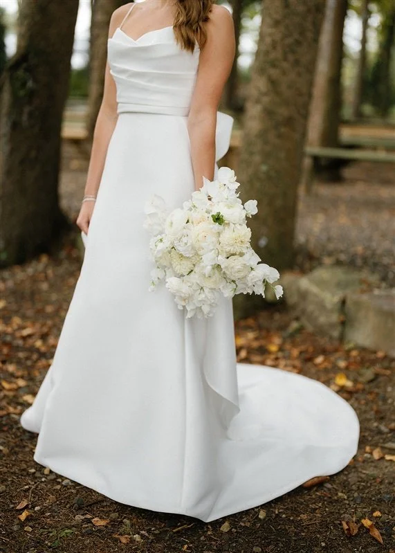 A bride in a strapless white wedding gown holding a bouquet of white flowers stands outdoors in a wooded area.