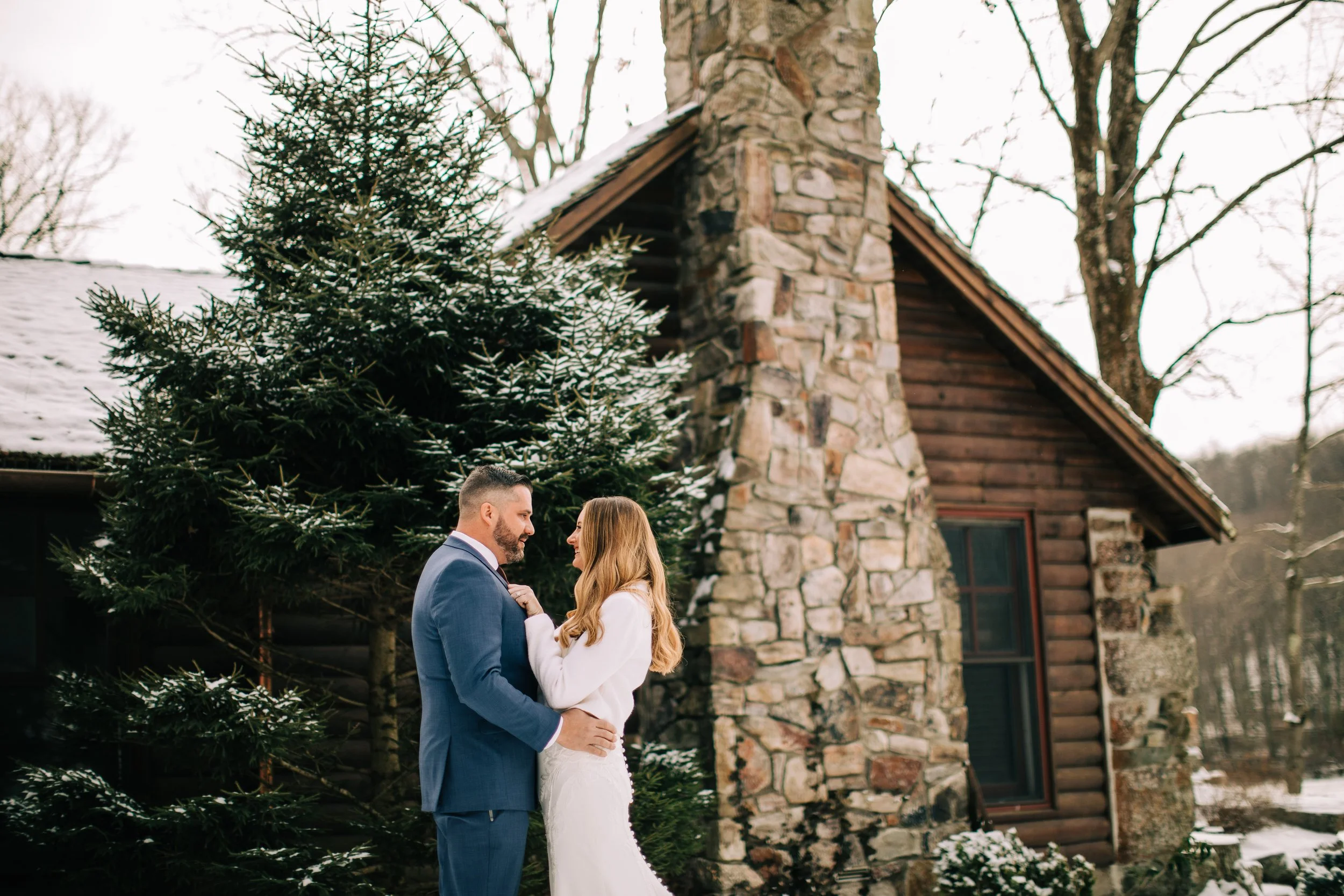A couple dressed in wedding attire standing close together outdoors in winter, near a large Christmas tree and a rustic stone and wood cabin.