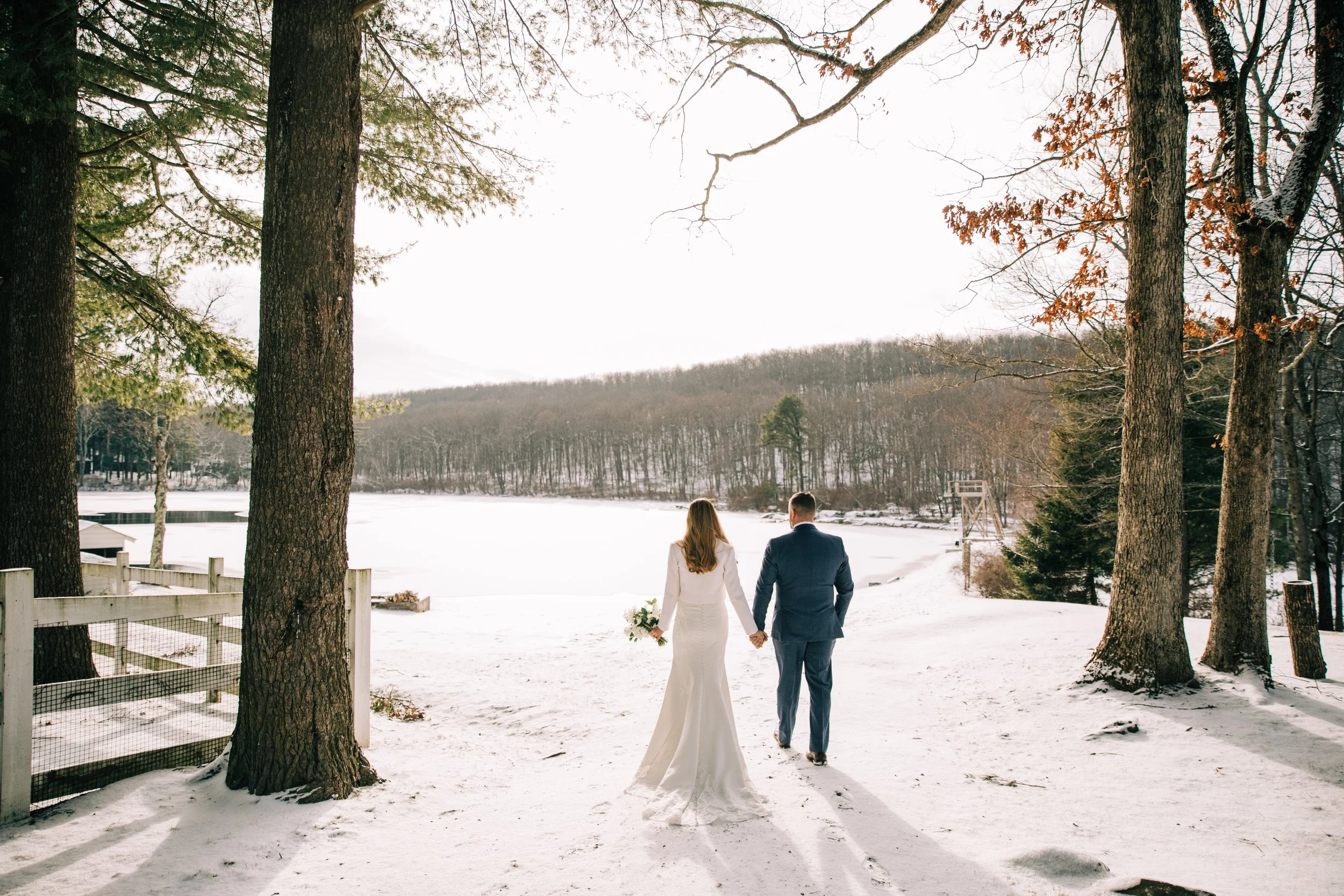 A bride and groom holding hands and walking on a snow-covered path in a winter landscape with a frozen lake, trees, and hills in the background.