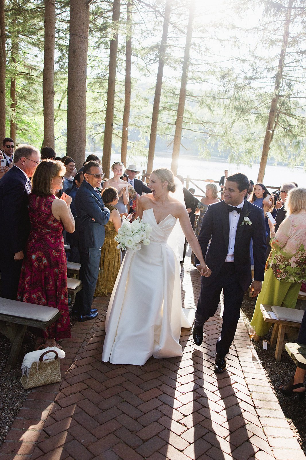 Bride and groom holding hands walking down an outdoor aisle at a wedding, surrounded by seated guests in a forested area near a lake.