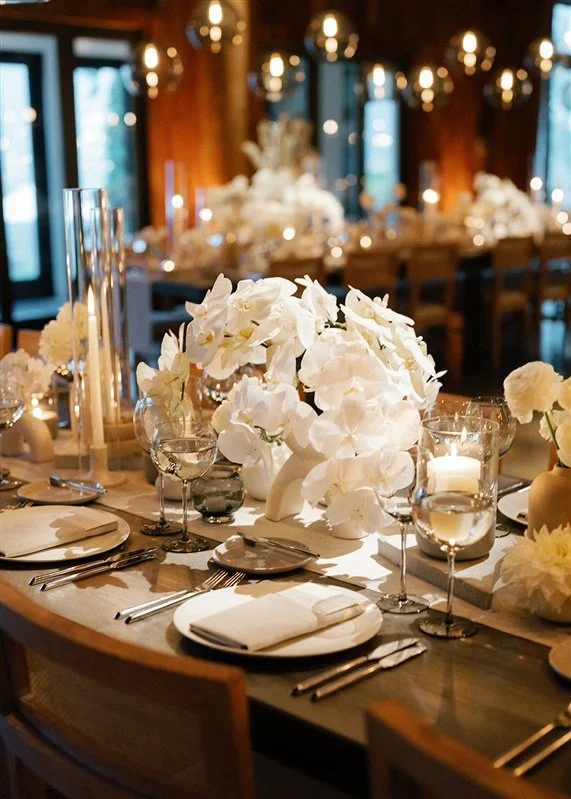 Elegant dining table with white floral centerpieces, candles, wine glasses, and place settings in a warmly lit, upscale restaurant or event space.