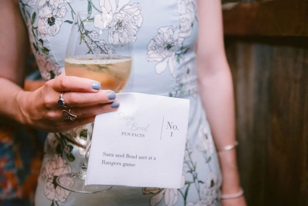 A woman with light blue nail polish holding a glass of white wine or champagne, with a printed card hanging from her hand that says "Sara & Brad," with some text about how they met and a number 1. She is wearing a floral dress, and the background app