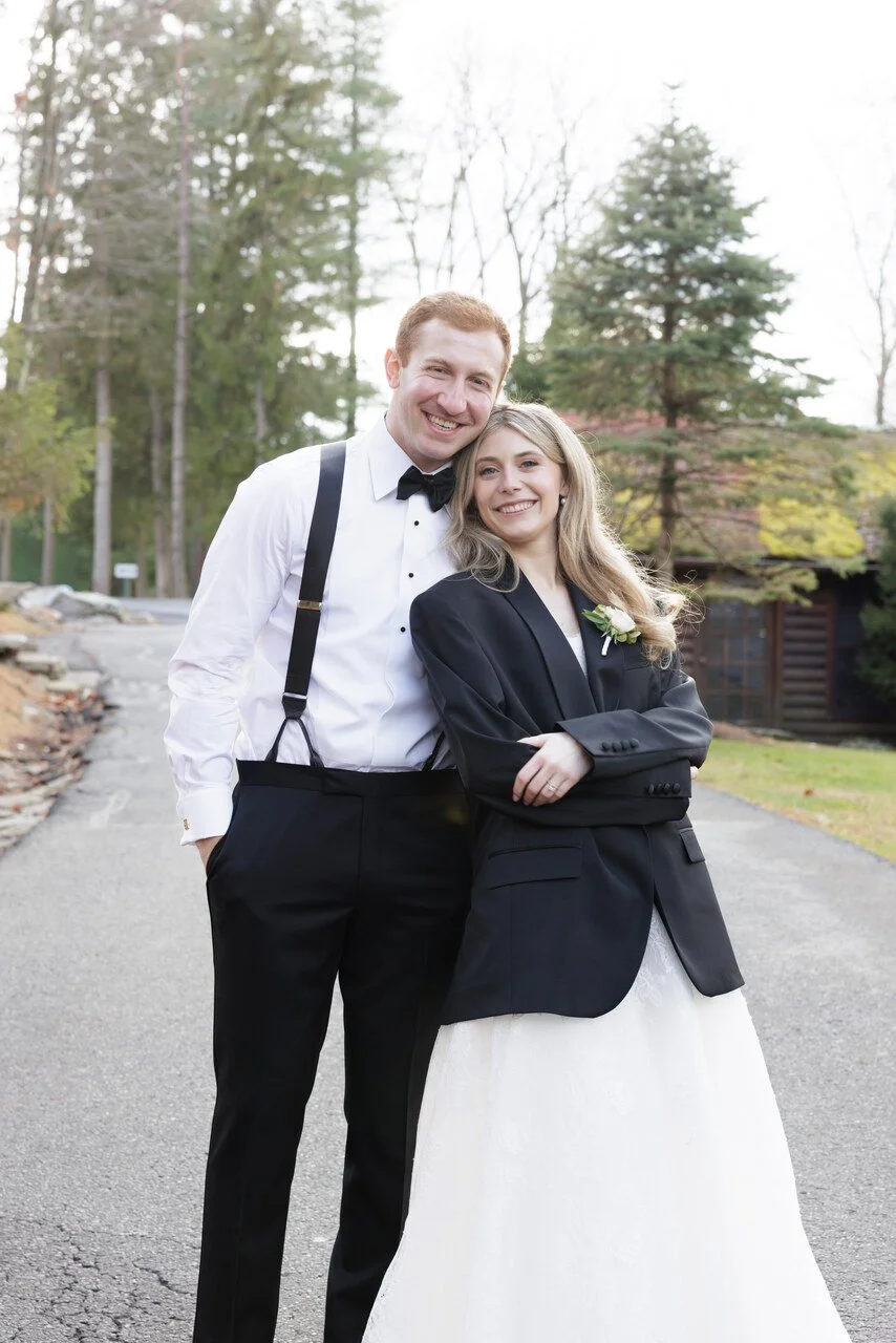 A couple dressed in wedding attire standing outdoors on a paved road, smiling and embracing, with trees and a house in the background.