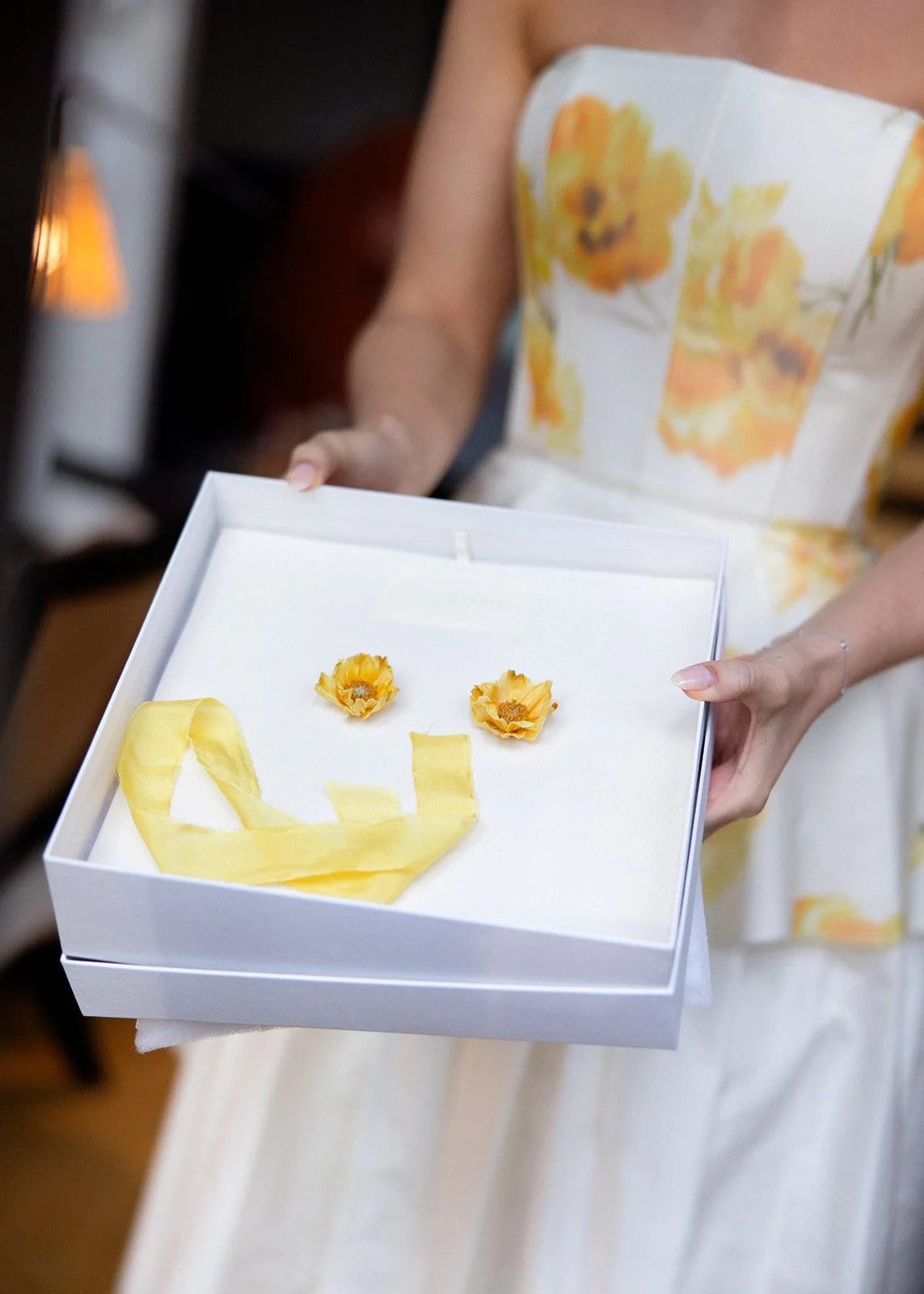 Person holding a white box with a yellow paper smiley face, made with two yellow flower-like objects for eyes and a yellow ribbon for the mouth, on a soft white surface inside the box.