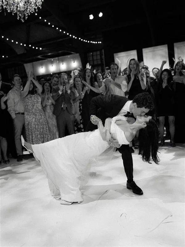 A couple is dancing at their wedding reception, the groom dips the bride while she leans back, and they kiss. Guests in the background are clapping and cheering, under string lights and a chandelier.