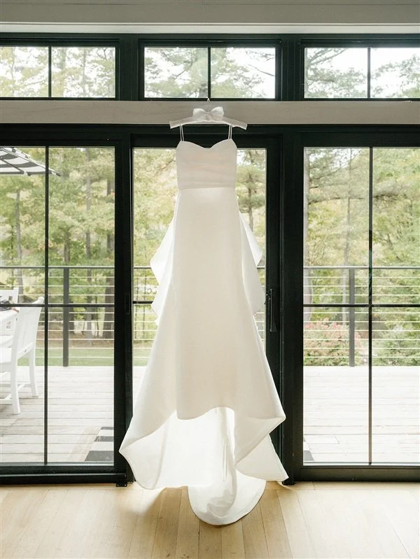 A white wedding dress hanging on a hook in front of a large glass door with a view of a wooden deck and trees outside.
