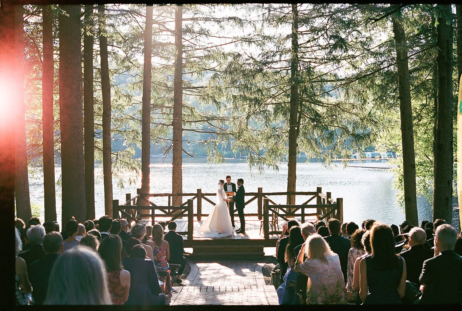 A wedding ceremony is taking place outdoors on a wooden platform near a lake, surrounded by tall pine trees, with guests seated and watching the event.