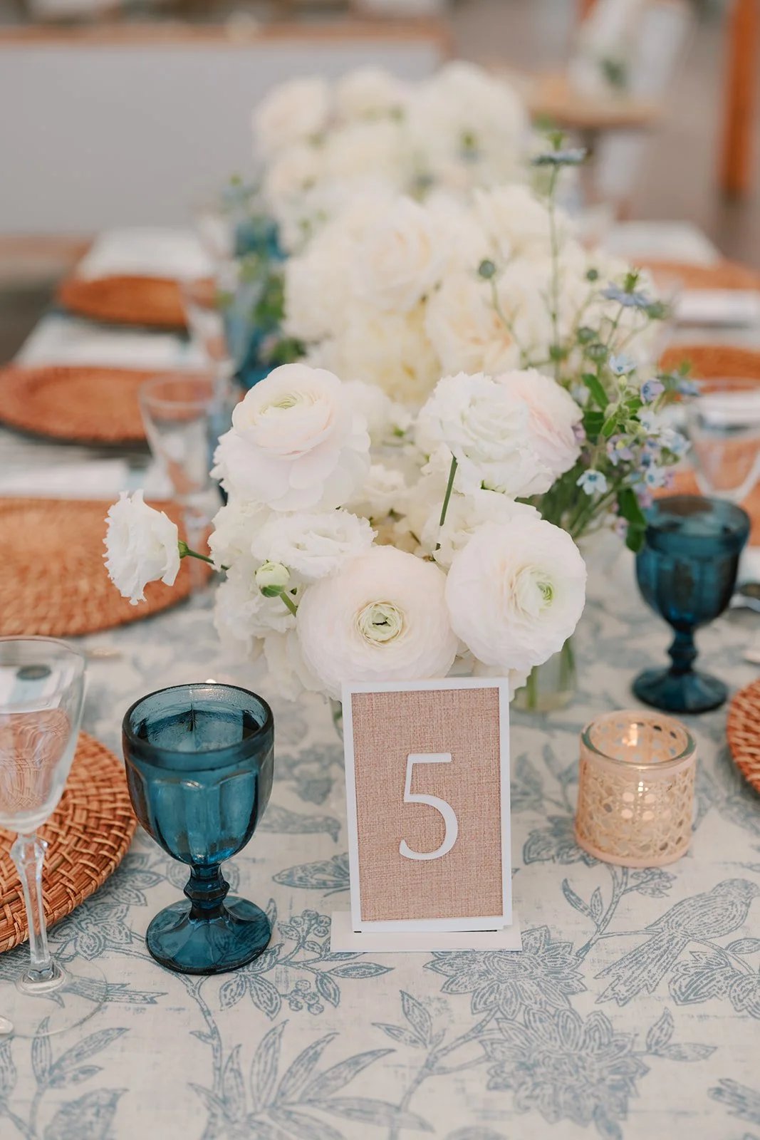 Table setting with a centerpiece of white flowers, blue glasses, and a table number '5' on a card.