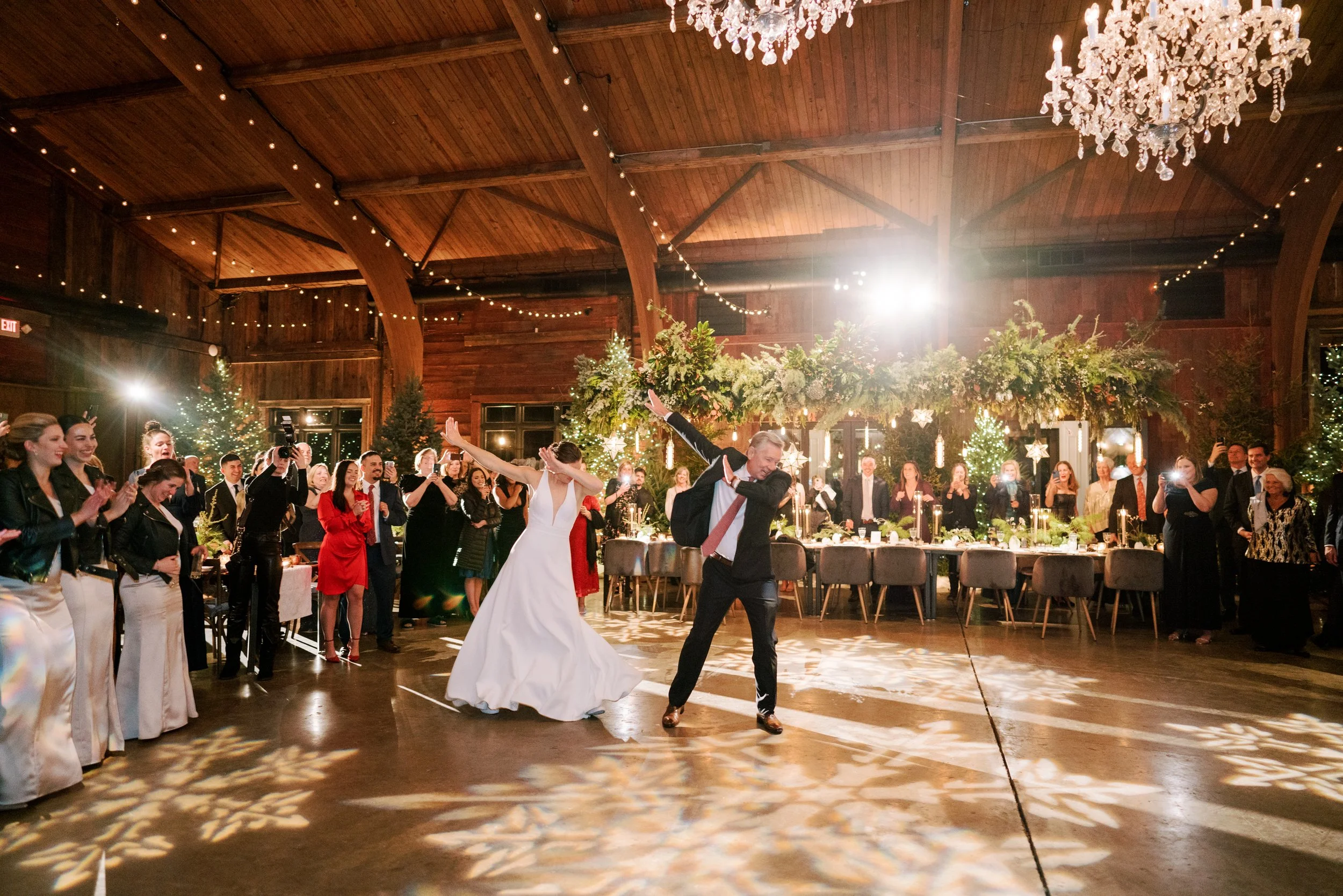 A bride and groom dancing at their wedding reception with guests gathered around, decorated with festive lights and trees, in a rustic wooden venue with chandeliers and holiday decor.