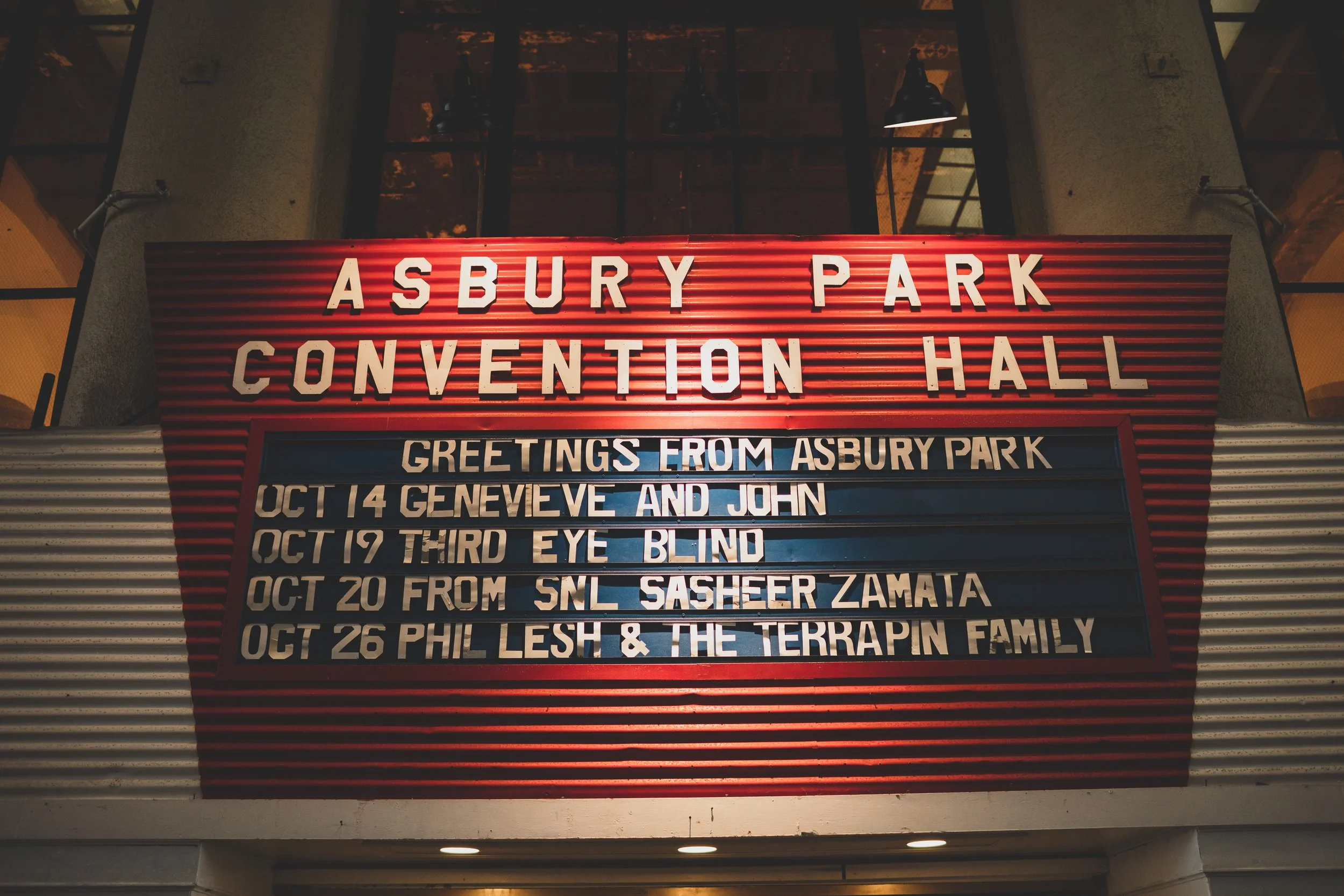 A sign outside the Asbury Park Convention Hall displaying event names and dates, including greetings from Asbury Park, with a red background and black lettering.