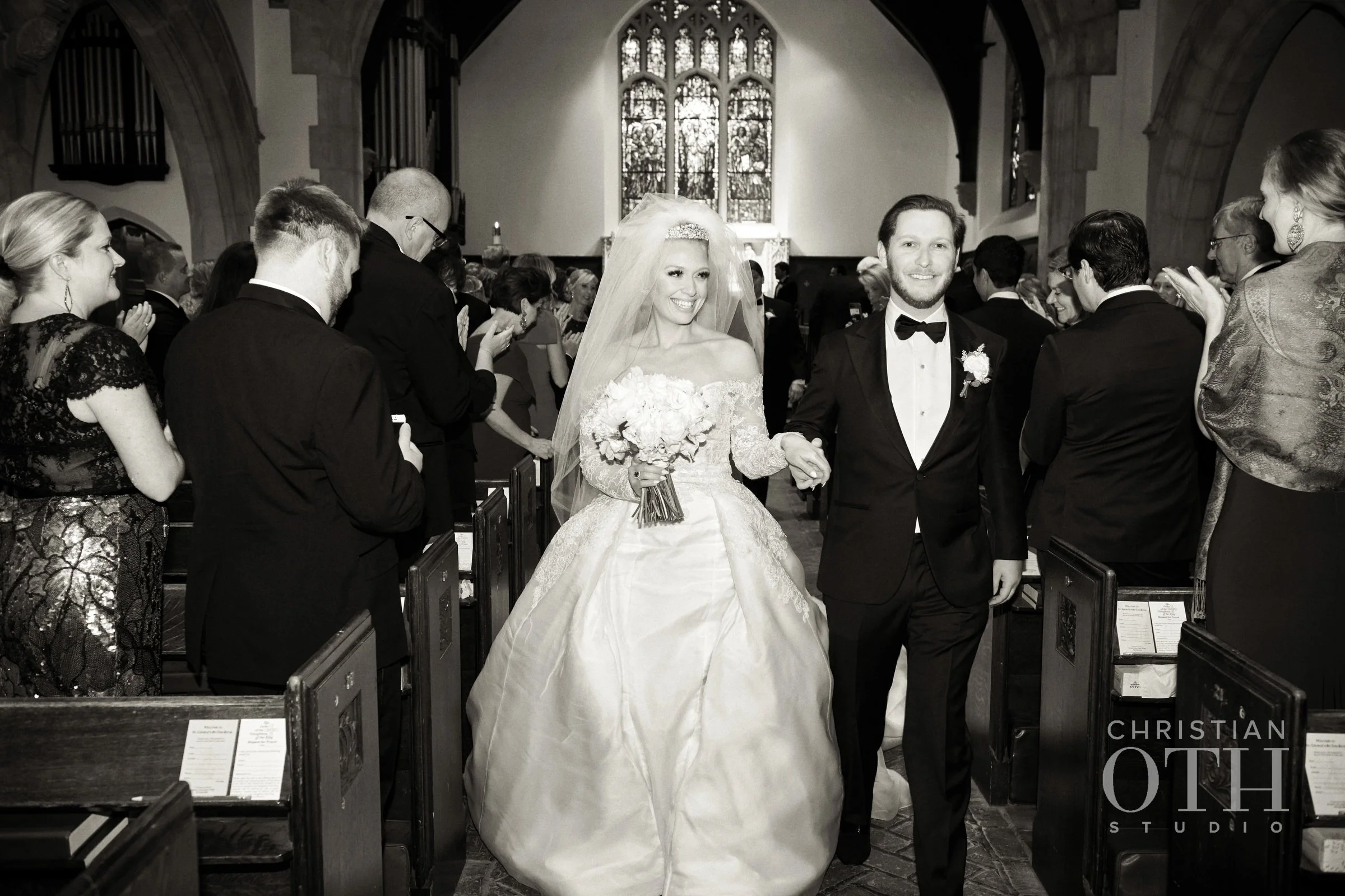 A bride and groom walking down the aisle of a church after their wedding ceremony, surrounded by guests who are clapping and smiling.