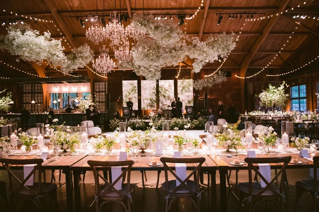Elegant wedding reception in a rustic wooden venue with floral decor, chandeliers, and string lights. Long dining tables adorned with white flowers, candles, and place settings, with a stage at the back featuring a bride and groom silhouette.