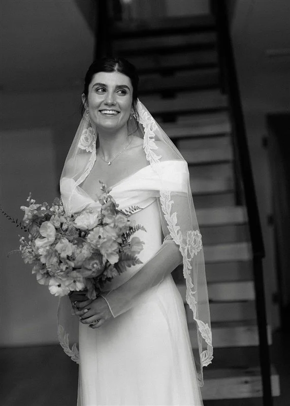 Black and white photo of a smiling bride in a wedding dress with a lace veil, holding a bouquet of flowers, standing indoors near a staircase.