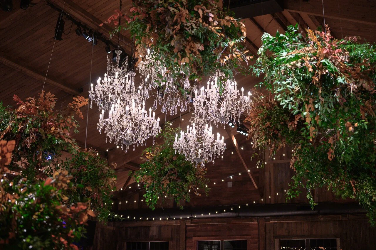 Chandeliers hanging from a wooden ceiling with greenery and string lights.