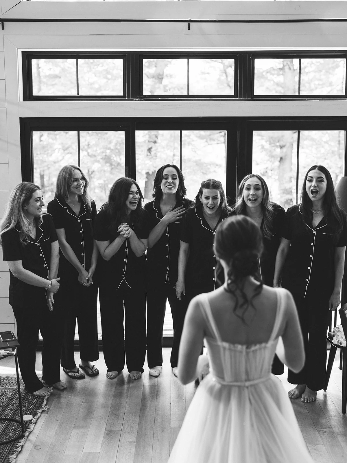 A bride with wavy hair in a white dress faces a group of seven women in matching pajamas, smiling and singing, inside a room with large windows showing trees outside.