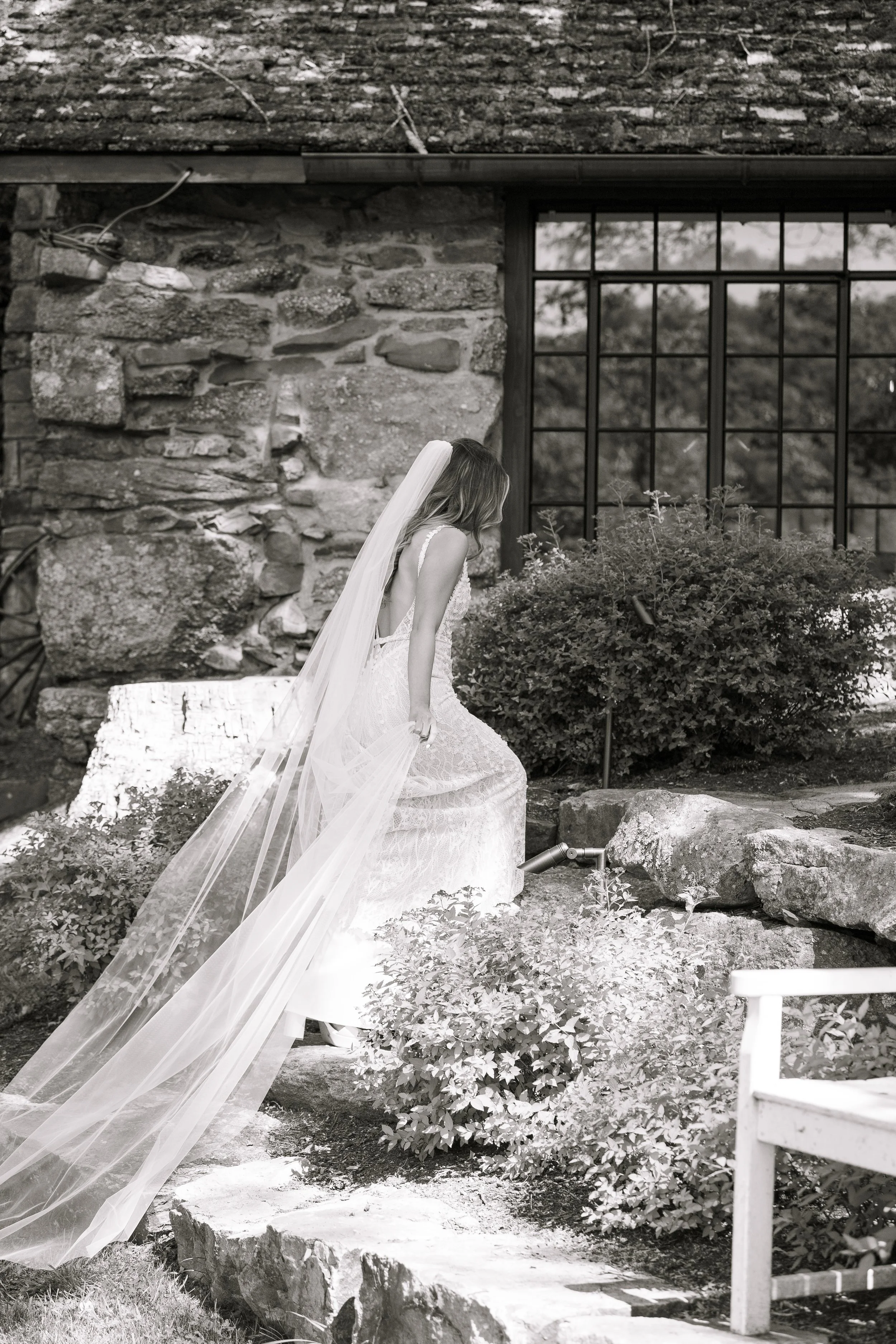 A bride in a wedding dress with a long veil, sitting on a rock in front of a rustic stone building with large window.