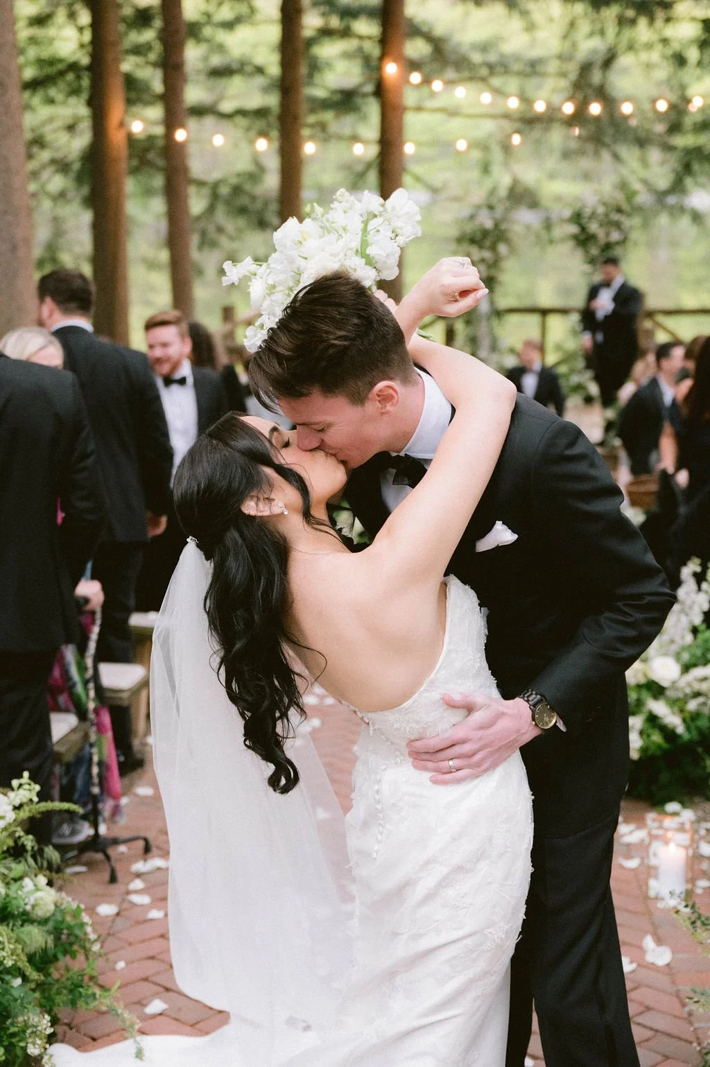 A newlywed couple sharing a kiss at their wedding reception inside a wooded venue with string lights hanging overhead.