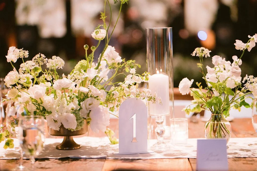 Floral centerpiece with white flowers on a wedding or event table, accompanied by a tall glass vase with a candle inside, a table number card, and other floral arrangements in glass vases.