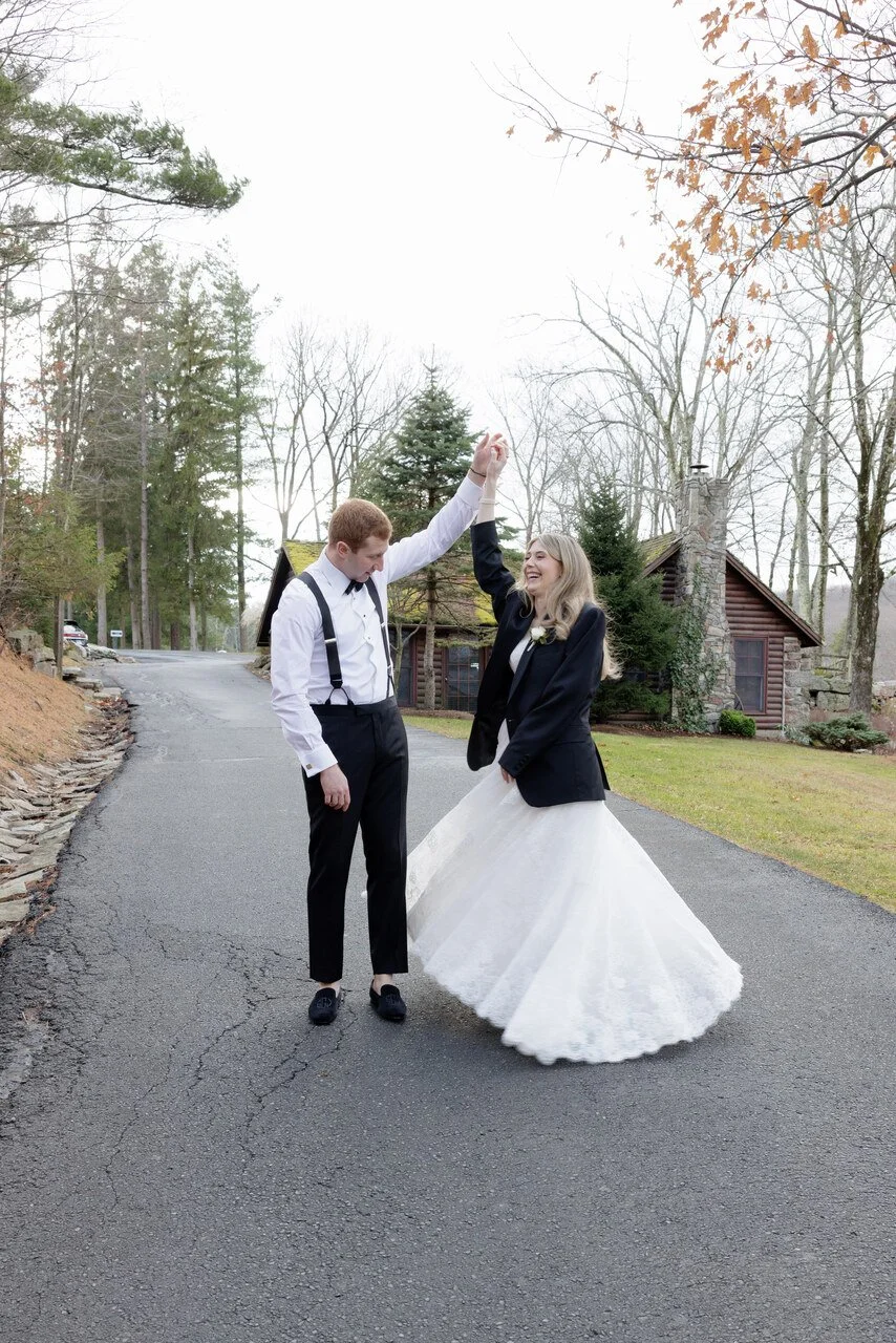 A bride and groom dancing outdoors on a paved road in front of a rustic house, with trees around them. The groom is wearing a white shirt with suspenders and black pants, while the bride is in a wedding dress with a black blazer. They are smiling and