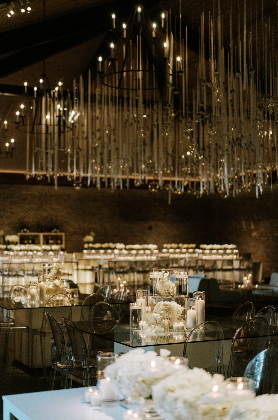 Elegant event space decorated with white flowers and candles, featuring a modern chandelier with numerous hanging candles, set for a celebration or gathering.