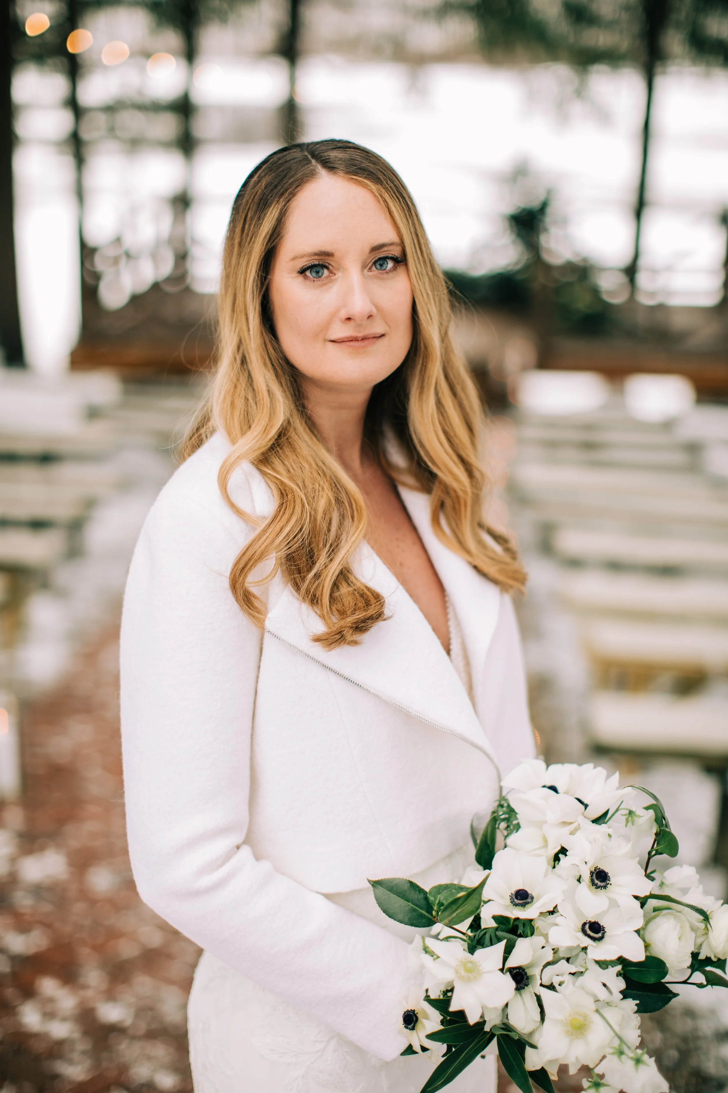 A woman with long blonde hair and blue eyes wearing a white coat, holding a bouquet of white flowers, standing outdoors near a lake with trees in the background.